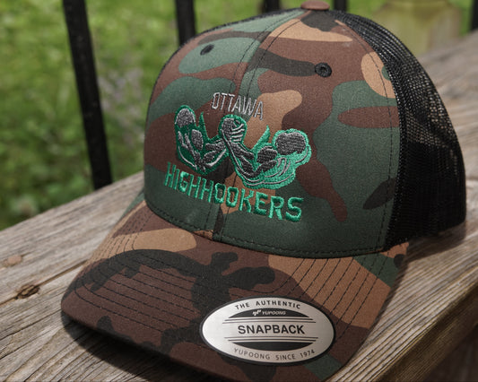 A camouflage snapback trucker hat with black mesh back panels, featuring a green embroidered 'Ottawa Highhookers' logo on the front. The hat is resting on a weathered wooden deck railing with a blurred green outdoor background.