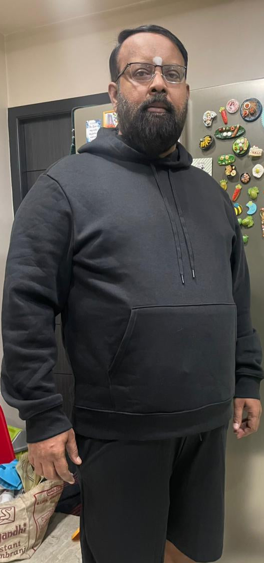 A man wearing a dark gray pullover hoodie and black shorts stands in a kitchen in front of a Samsung refrigerator.