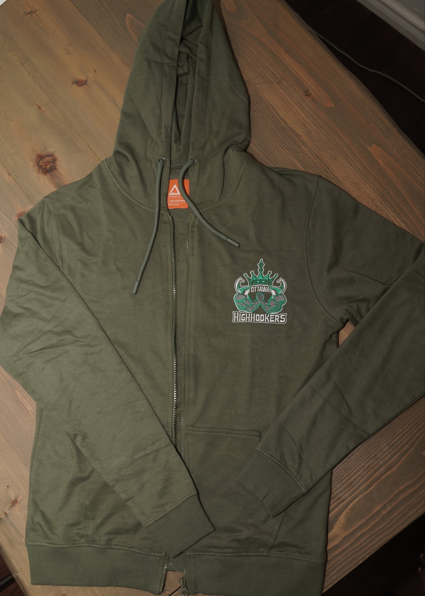 An olive green zip-up hoodie laid flat on a wooden table, featuring a small graphic on the left chest \