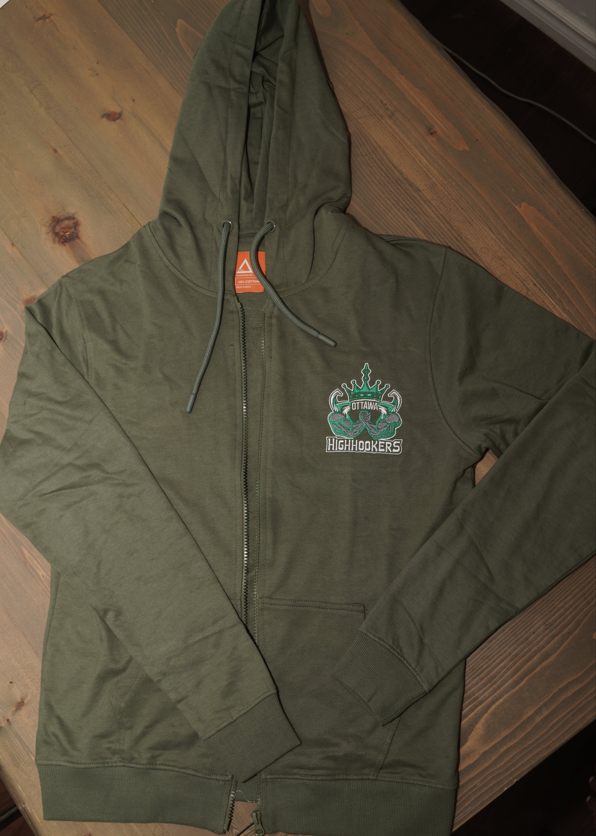 An olive green zip-up hoodie laid flat on a wooden table, featuring a small graphic on the left chest \