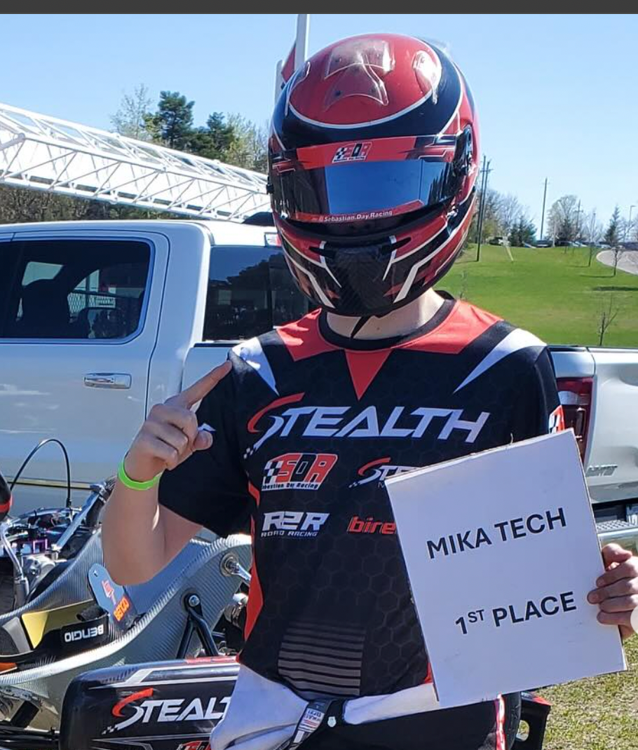 Person in racing gear holding a '1st Place' sign with a vehicle and track in the background