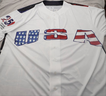 A white short-sleeve button-up baseball jersey laid flat on a bed, featuring large 'USA' lettering across the chest in an American flag pattern and a small patch on the right sleeve.