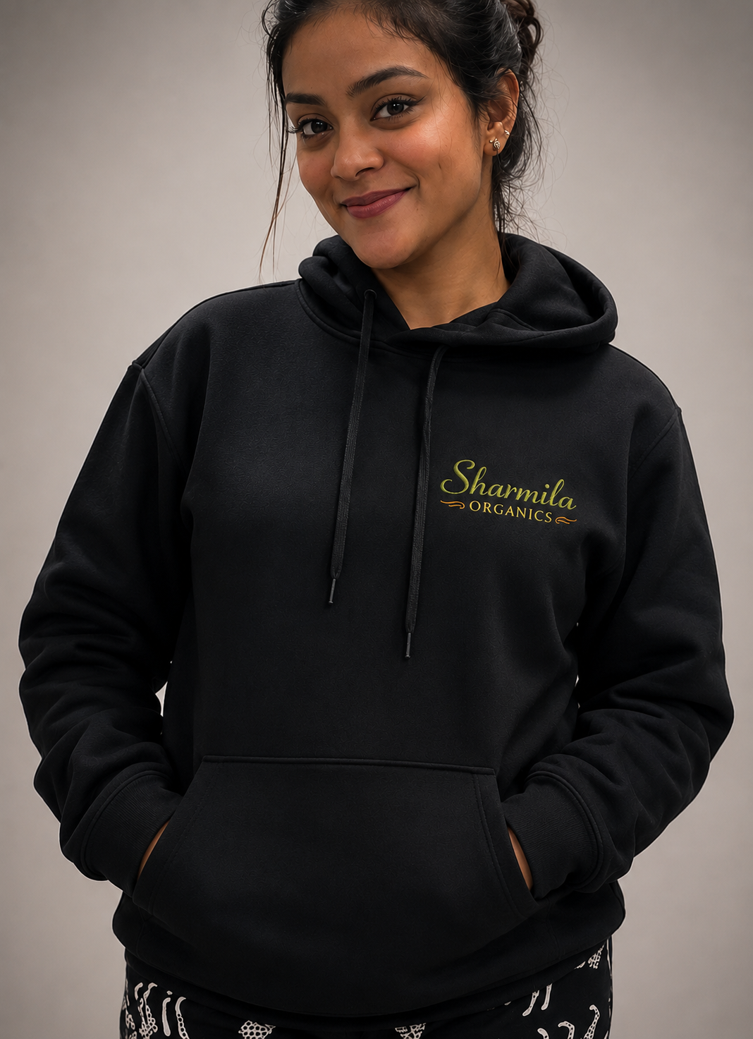A woman wearing a black pullover hoodie with 'Sharmila Organics' printed in gold on the left chest, posed against a neutral gray background.