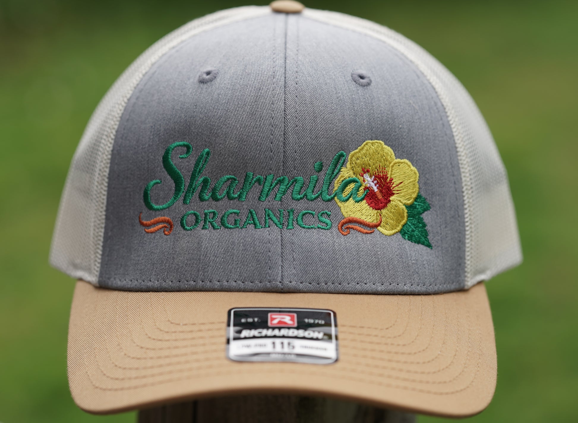 A close-up of a gray and beige trucker hat with 'Sharmila Organics' embroidered on the front alongside a yellow hibiscus flower. The hat has a tan visor and a green, red, and orange embroidered logo.