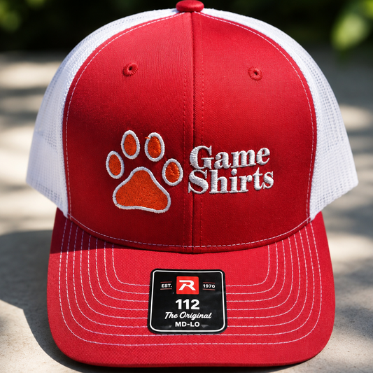 A red and white trucker hat with a paw print and 'Game Shirts' logo on the front, displayed on a stone surface with a blurred outdoor background.